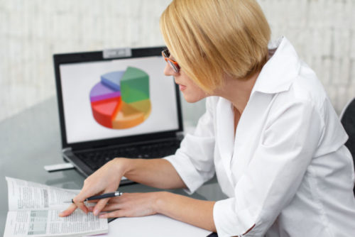 A woman looking over financial documents, while a chart is displayed on her computer.