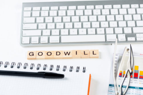 Letter tiles spell out "goodwill" and are surrounded by a computer keyboard, a pen and notepad, and eyeglasses sitting on top of a financial chart document.