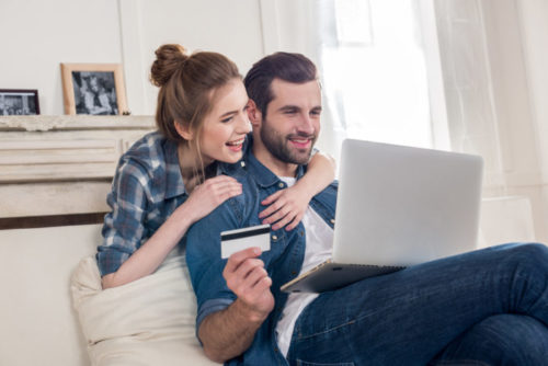 The man of a smiling couple who is shopping online holds up a credit card.