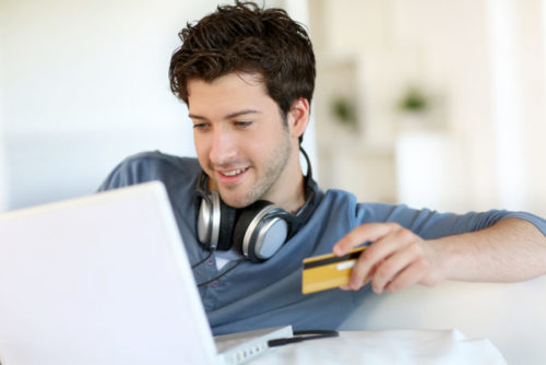 A smiling man with headphones on using his card to make a purchase on his laptop.
