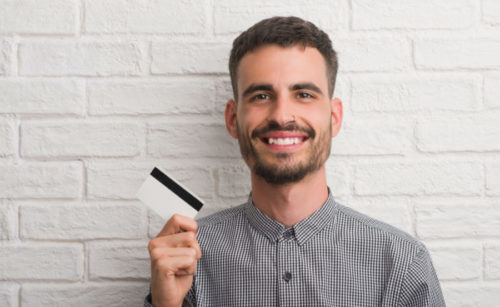 A smiling man stands in front of a white brick wall while holding his credit card.