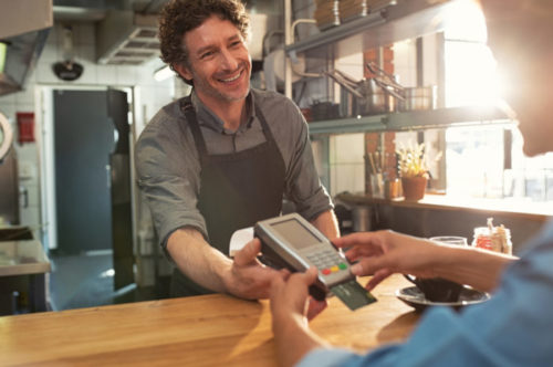 A smiling barista handing a customer a payment terminal to pay for her coffee with a credit card.