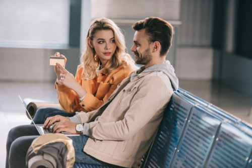 The woman of a smiling couple in an airport points at her credit card while looking at her husband.