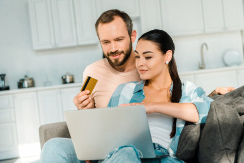 The man of a smiling couple holds a credit card while the woman uses a laptop.