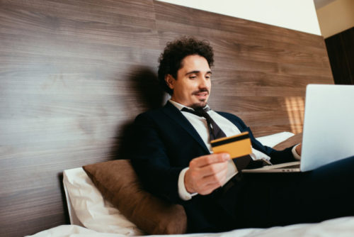 A businessman sitting on a hotel bed using his laptop and holding a credit card.