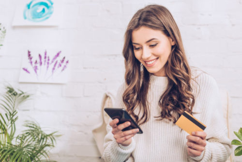 A smiling woman shops on her phone while holding her credit card.