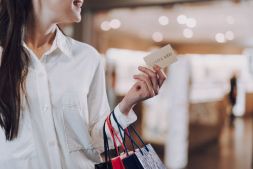 A smiling woman with shopping bags on her arm holds her credit card up.