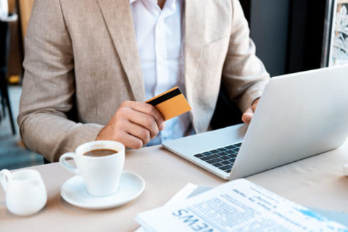 A person in business attire working on their laptop readies his credit card to pay at a cafe.