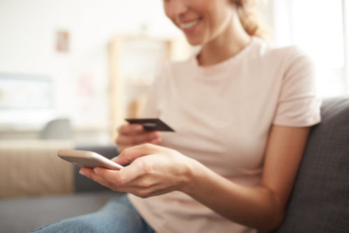 A smiling woman, sitting on her couch, holding her phone and her credit card.