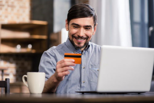 A person smiles while looking at their credit card while working on their laptop in a cafe.