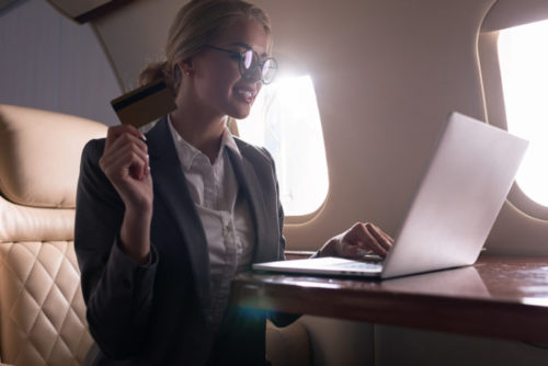 A person in business attire works on a laptop on a plane while holding a credit card.