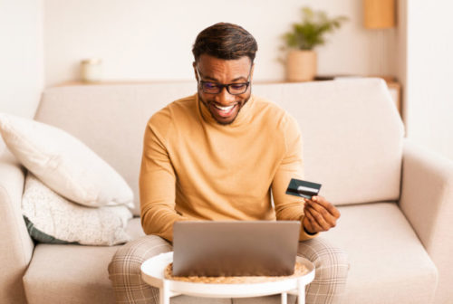 A man smiles while using his laptop and holding his credit card.