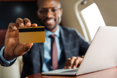 A person in business attire working on a laptop holds up a credit card and smiles while on an airplane.