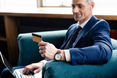 A person in business attire uses a laptop while holding up his credit card and smiling.
