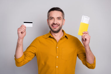 A smiling man holds plane tickets in one hand and a credit card in the other.