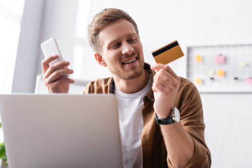 A person working on their laptop, smiling while holding their phone and credit card.