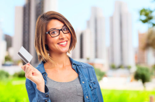 A smiling woman holds up her credit card outside.