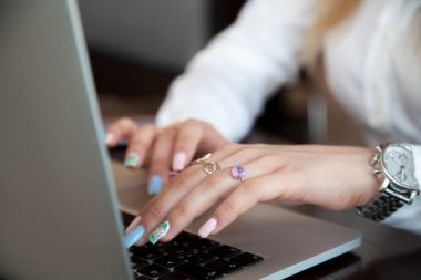 A woman with painted nails and rings on her fingers types on a laptop.