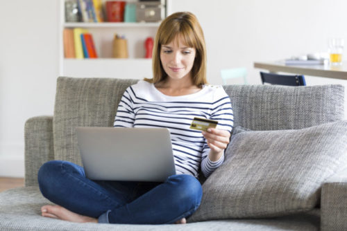 A person sitting on their couch, using their laptop to shop online while holding their credit card.