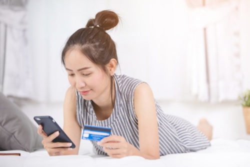 A person lying down on a bed, shopping on her phone while holding a credit card.