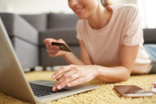 A smiling person lying down, typing on a laptop while holding a credit card.
