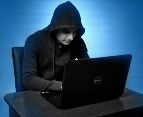 An internet scammer, dressed in all black, sits at a laptop.