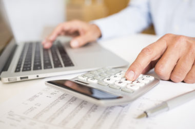 A person typing on a computer while tallying financial figures on a calculator.