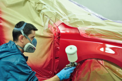 An auto professional painting a car with a sprayer while wearing a respirator mask.
