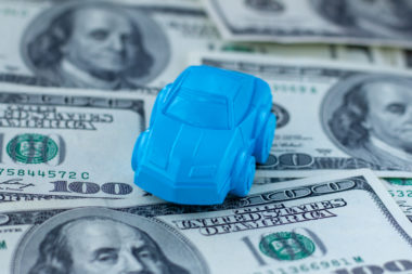 A tiny blue model car sits on top of loan money in the form of $100 bills.
