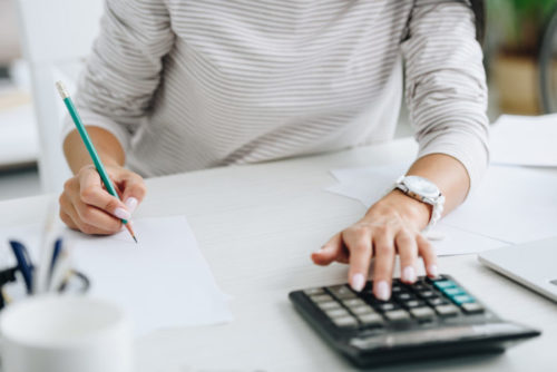 Someone sits at a desk and uses a pencil, paper, and calculator to work out calculations by hand.