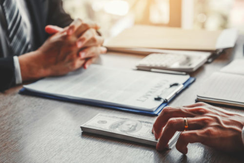 A loan officer sits down with a contract while their client picks up a stack of cash.