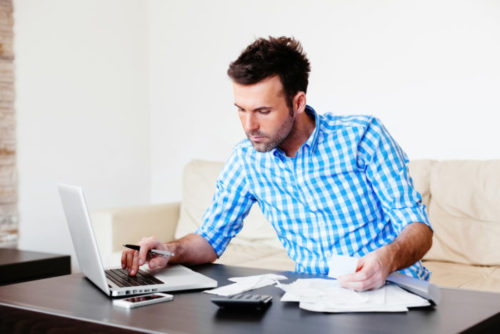 A person surrounded by a phone, calculator, a laptop, and various invoices in an effort to pay their bills.