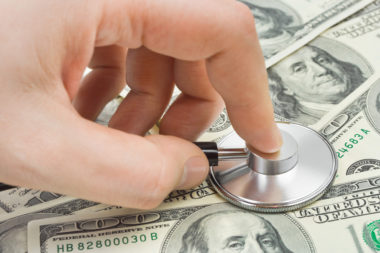 A closeup of a persons hand placing a stethoscope on top of various $100 bills.