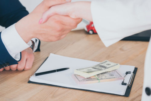 Money sits on top of a loan contract, which is under a loan officer and a client shaking hands.