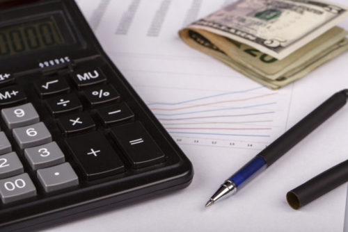 A calculator, a pen, and some cash are placed on top of financial documents.