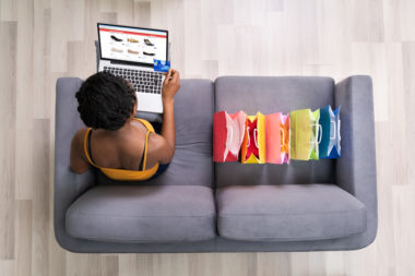 A person sits on their couch, holding their credit card and shopping online with shopping bags next to them.