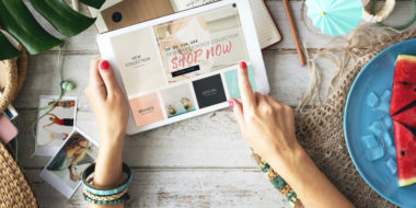 A person uses a tablet that displays a screen that reads "shop now" at their desk, which contains plants and food on it.