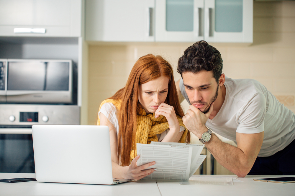 A couple look worried as the examine financial documents in their kitchen, in front of a laptop.