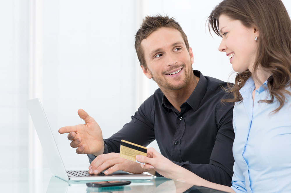 A person holds a credit card while another person speaks to them while typing on a laptop.