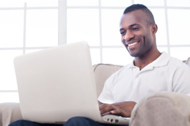 A person smiles while using a laptop on their couch.