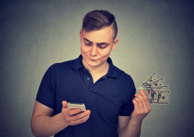 A person holds their phone on one hand while holding a fan of $100 bills in the other.