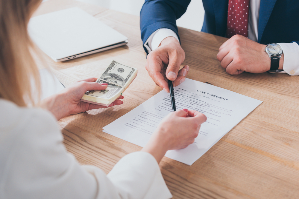 A person holds a stack of money while a loan officer hands them a pen to sign a contract.