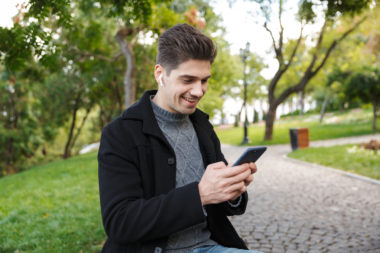 A person smiles as they read their phone in a park.