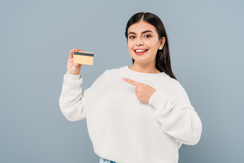 A person in a white sweater holding a credit card while pointing to it.