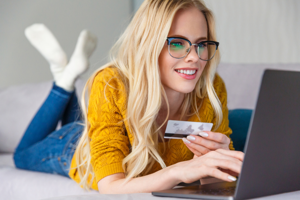 A person wearing a yellow shirt lies on their stomach while holding a credit card and shopping on their laptop.