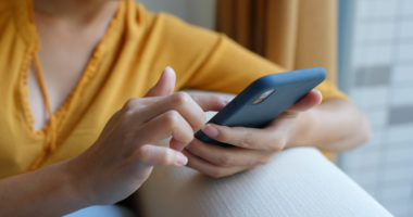 A person wearing a yellow shirt sits on their couch, leaning against the back of it, using their phone.