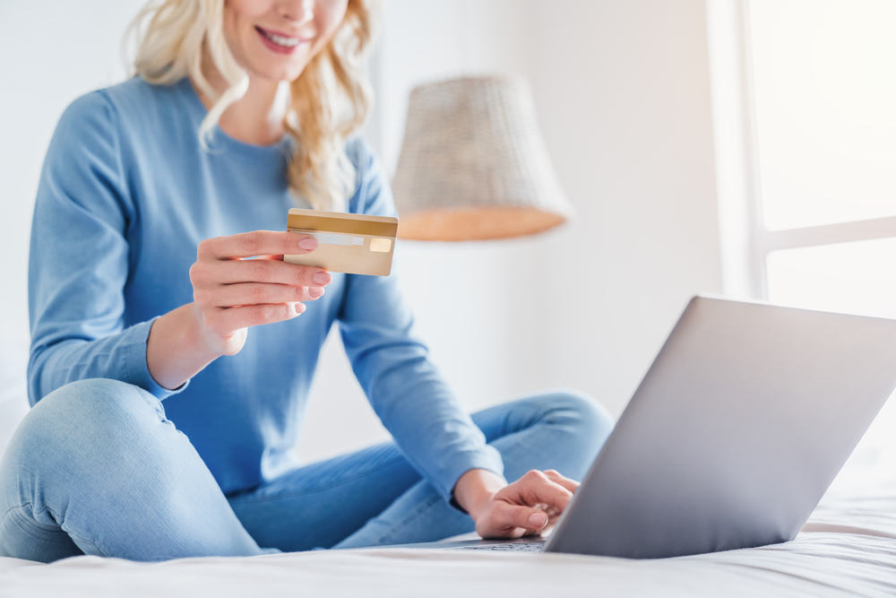 A person sitting on their bed, holding a credit card as they shop on their laptop.