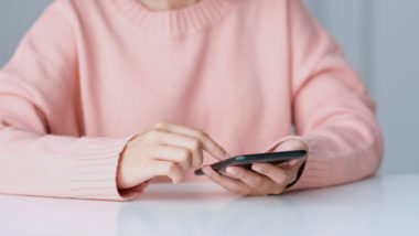 A person in a pink sweater sitting down at a table, using their phone.